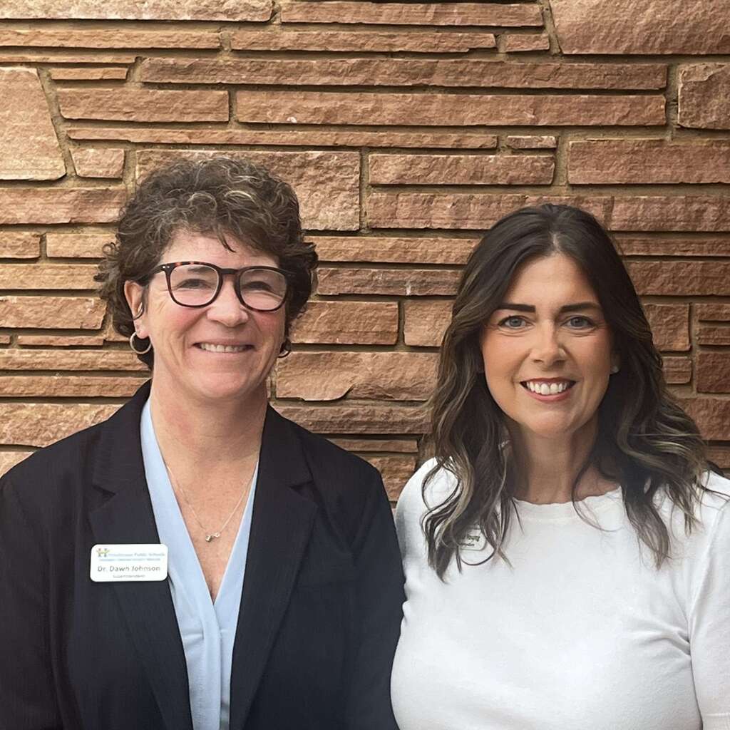 Two women pose together, smiling, in front of a brick wall.