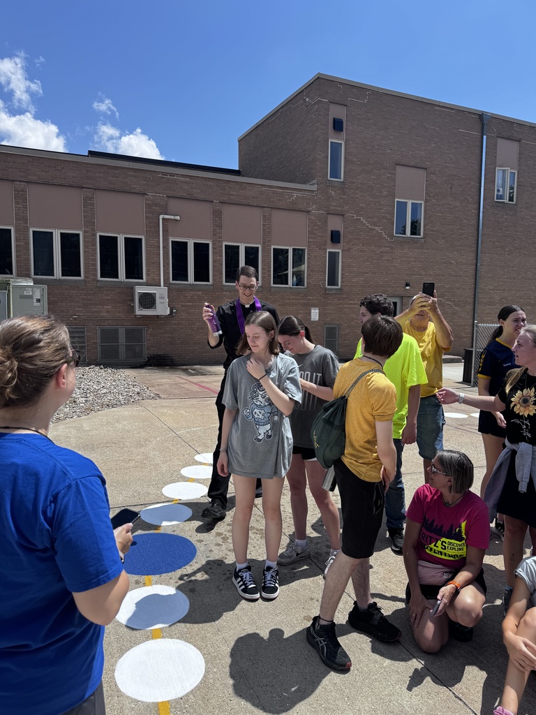 A priest blessings students that are standing a giant painted rosary