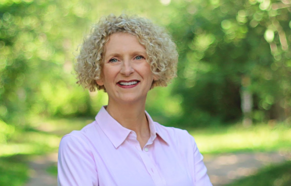woman standing with her arms folded, wearing a soft pink shirt and standing in front of green grass and trees in the background.