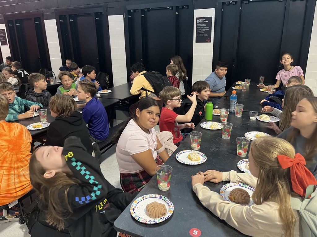 A group of children sitting at a long table, eating and drinking from plates and cups.