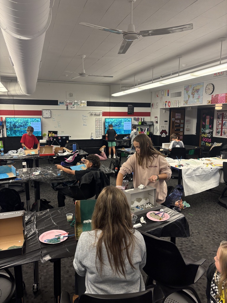 An art class with several students seated at tables; one student stands and works at the front.