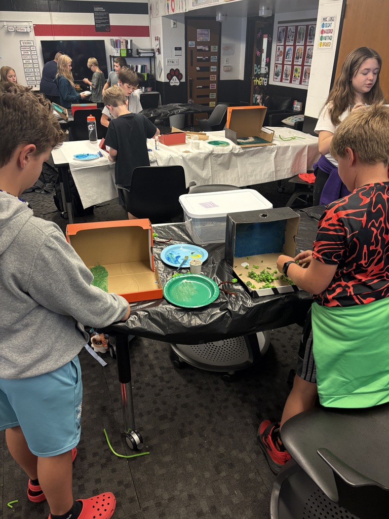 Kids in a classroom setting work on pizza boxes, one with a green plate and another with a pizza cutter.
