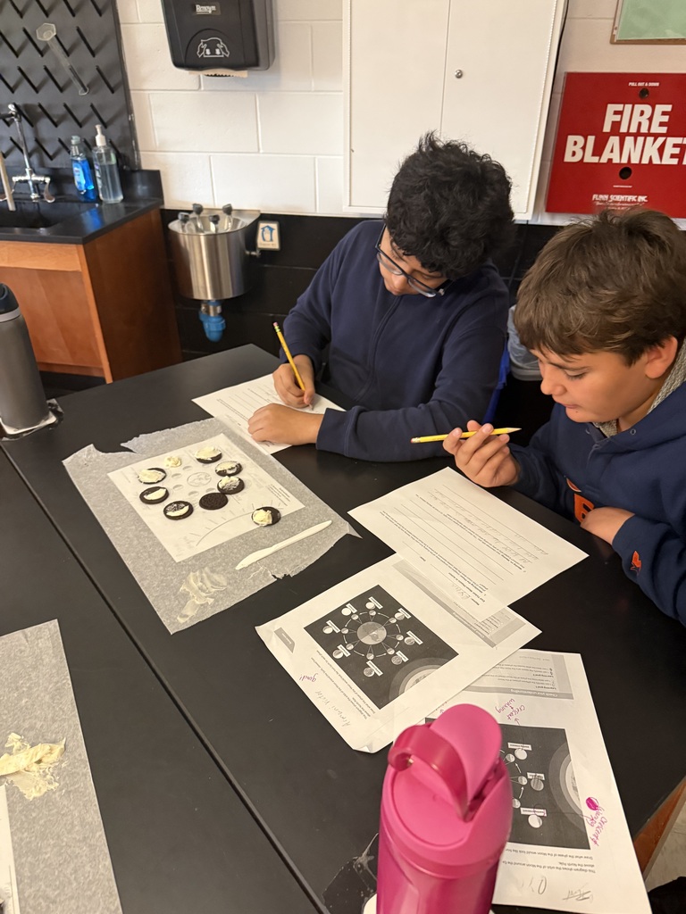 Two children sit at a table with papers and pencils, focusing on a worksheet. A trash can is nearby.