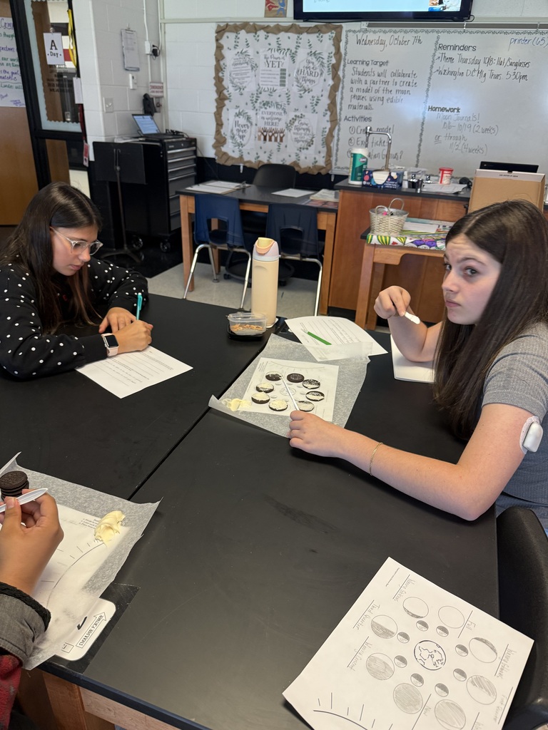 Two students work at a table with papers, pens, and a container. One holds a pen. Behind them, a whiteboard, desks, and chairs are visible.