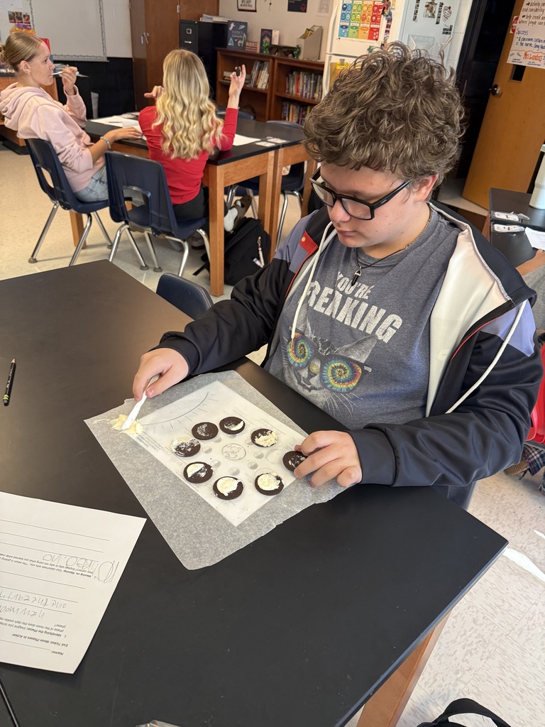 A student wearing glasses sits at a table arranging black and white cookies on a baking mat.