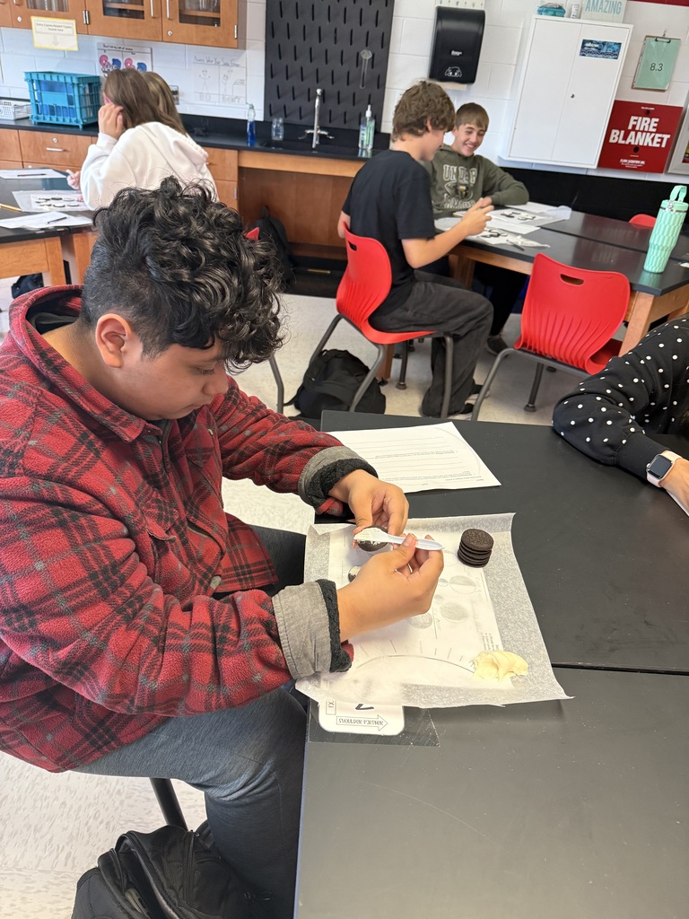 A student in a plaid shirt sits at a table, engaged in a lab activity with various tools and papers.
