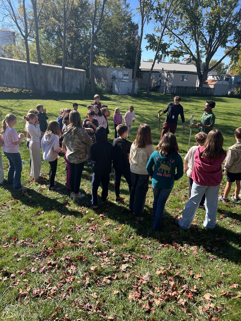 Students gather in an outdoor grassy area, with two adults standing in the center of the group.