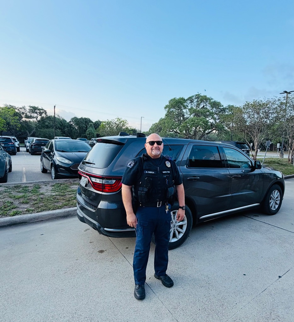 Police officer stands in front of black SUV, smiling, in parking lot with cars and trees.