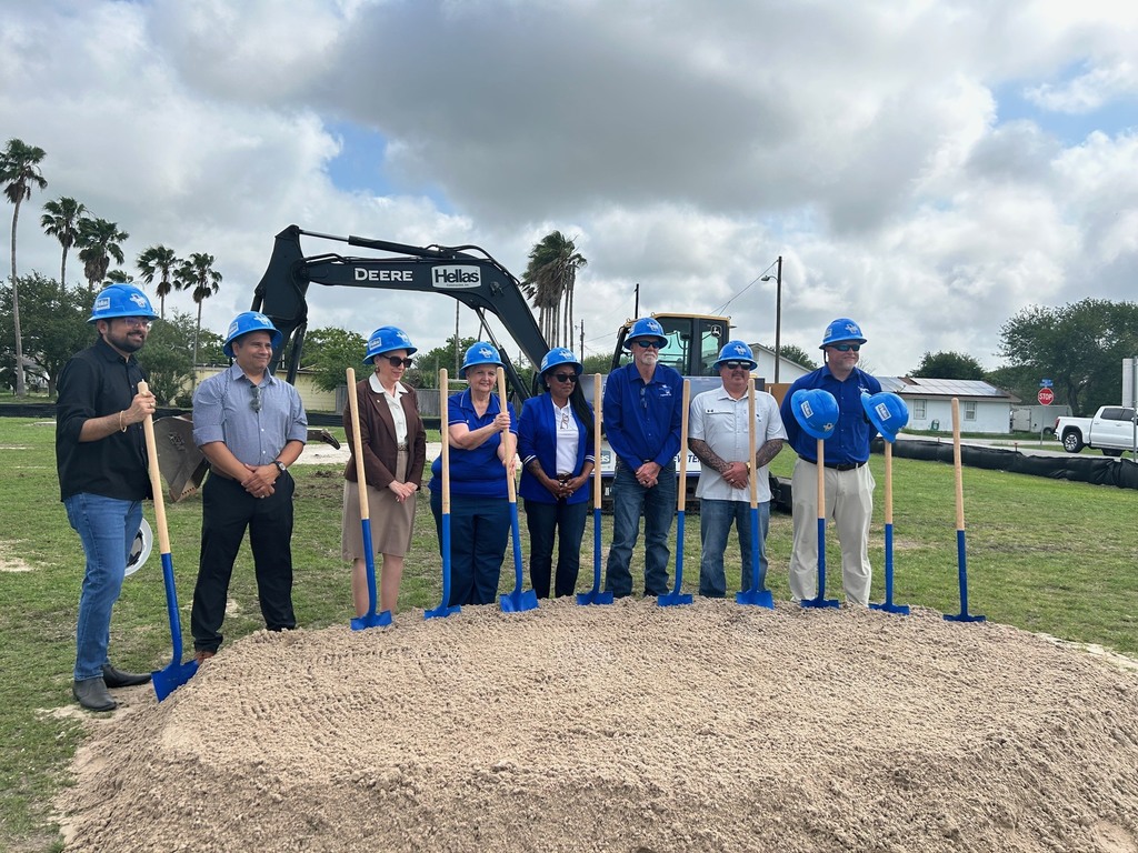 A group of people in blue helmets stands in front of a pile of dirt, holding shovels. A large machinery vehicle is behind them.