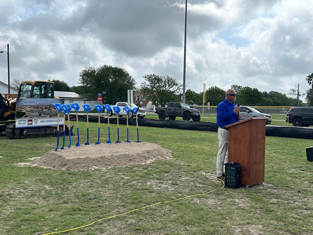 A man in a blue shirt stands at a podium in a field with blue construction markers.