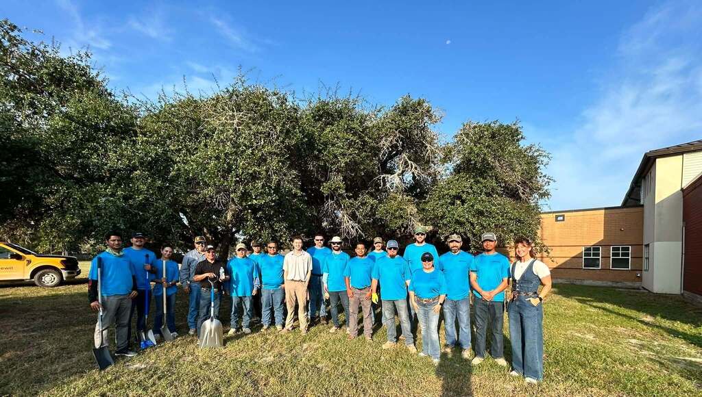 Group of people in blue shirts and jeans standing in a field, with trees and a building in the background.