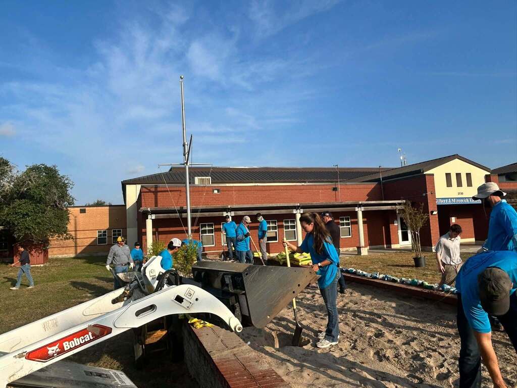 People in blue shirts work on a construction site with machinery. Behind them, a building has a brick facade and a flagpole.