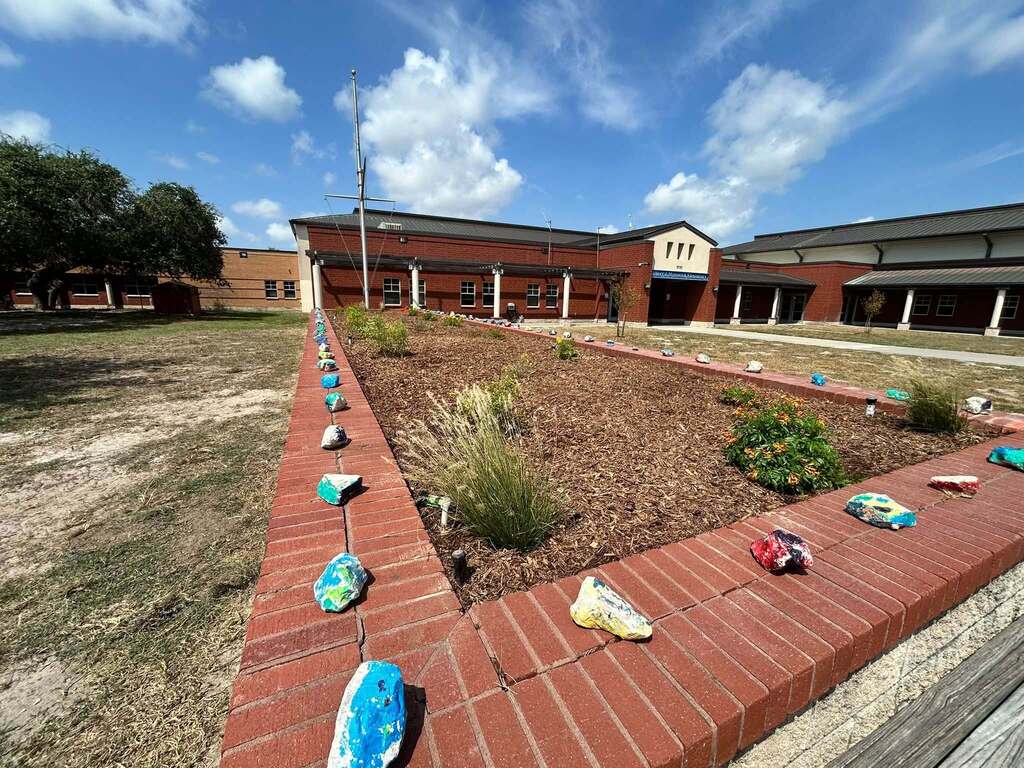 Brick walkway surrounds a garden area with plants and trees. A large building with a tower stands behind.