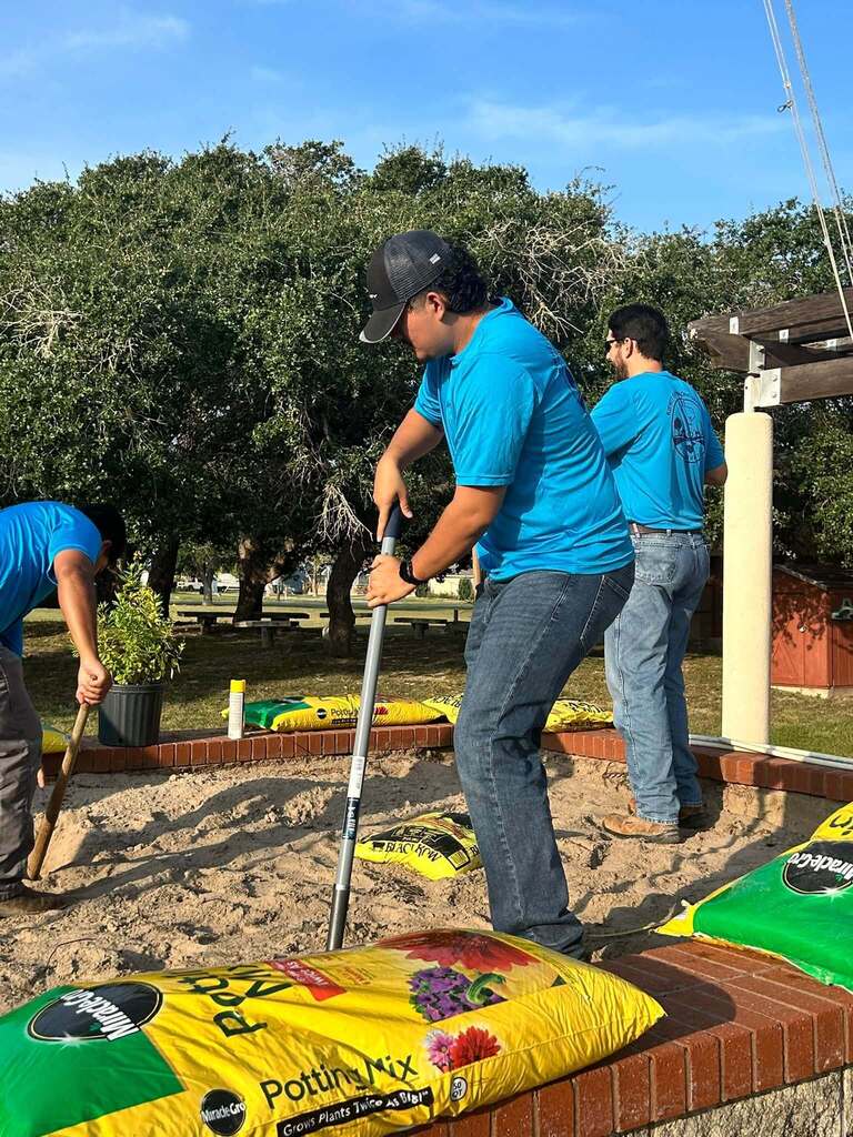 Three men in blue shirts and jeans using shovels and tools in a sandy area, with bags of potting mix.