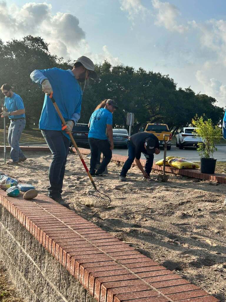 People in blue shirts and hats are working on a sandy area with tools, near a brick wall.