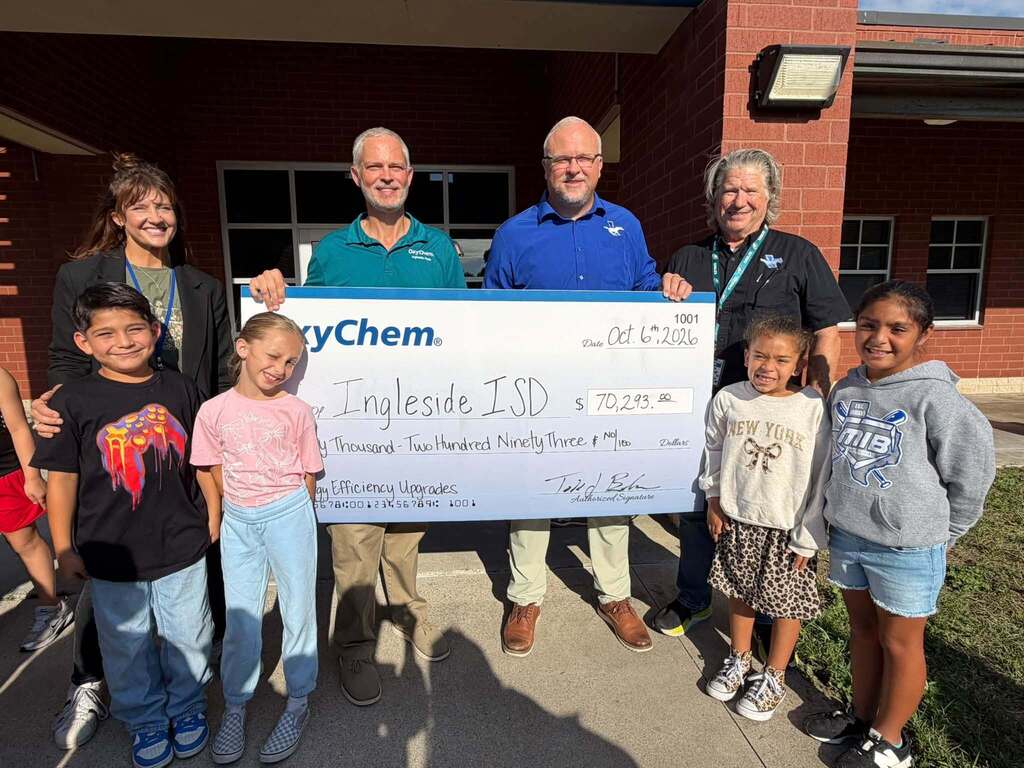 People stand in a group, smiling, in front of a brick building. Adults hold a large check. Children wear casual clothing.