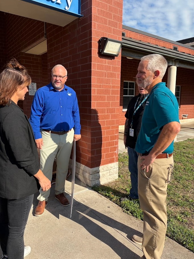 Four adults converse near a building. Two wear light blue shirts. Another wears a black shirt. A man uses a cane.