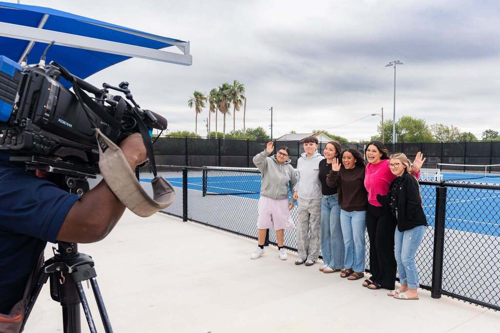 People posing for a photo by a tennis court, one person holding a camera on a tripod.