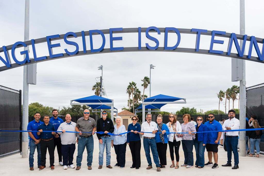People standing at a ribbon-cutting ceremony, smiling, under an arch sign with blue trim and palm trees in the background.