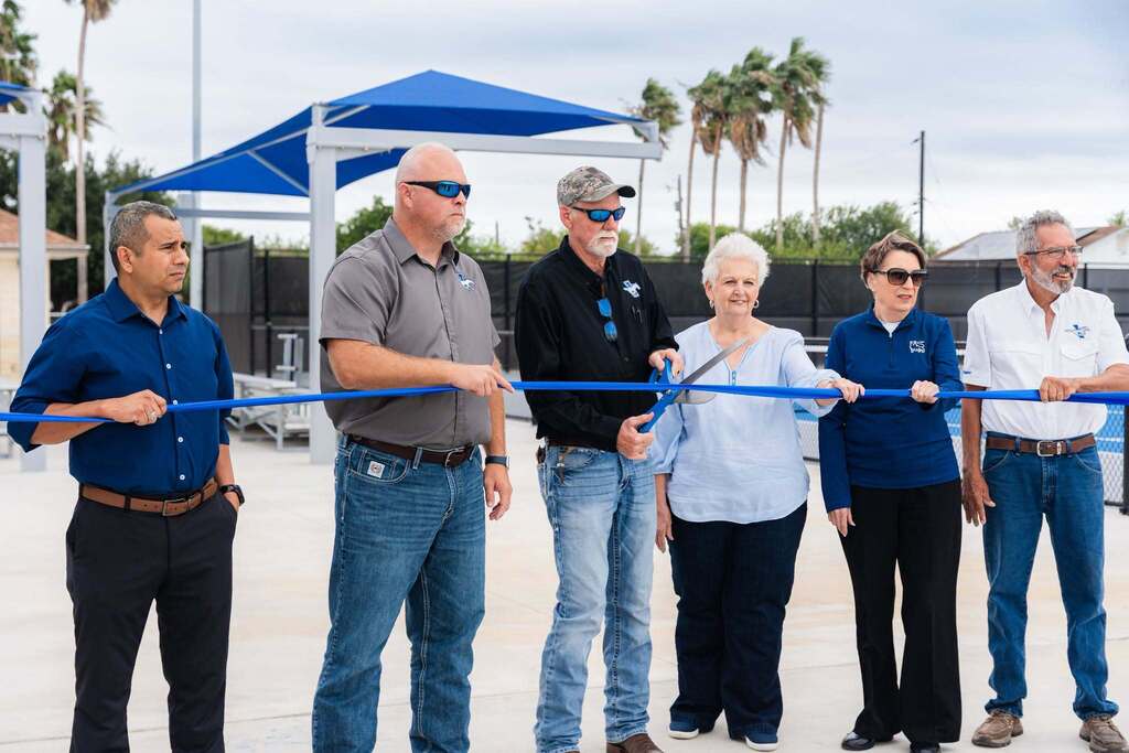 Adults stand in a line, holding a ribbon, ready to cut it. Trees and a blue canopy are in the background.