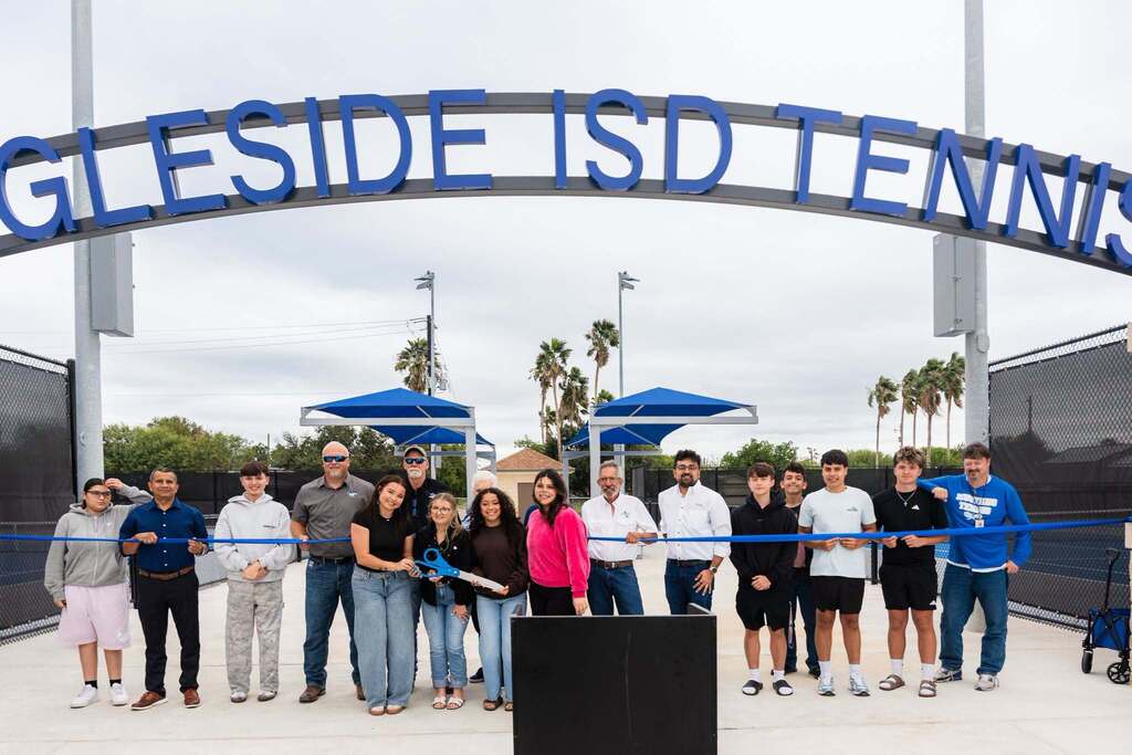 People stand in front of a tennis court entrance with a ribbon, smiling. A sign above reads "Gleside ISD Tennis."