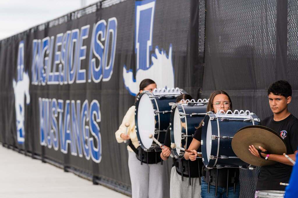 Three students holding drums stand near a wall with large text reading "CELESTE SD MUSTANGS".