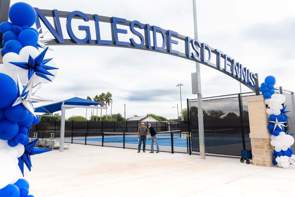 Entrance arch for Angleside Tennis Courts, adorned with blue and white balloons. Two people stand by a black fence, with tennis court in background.