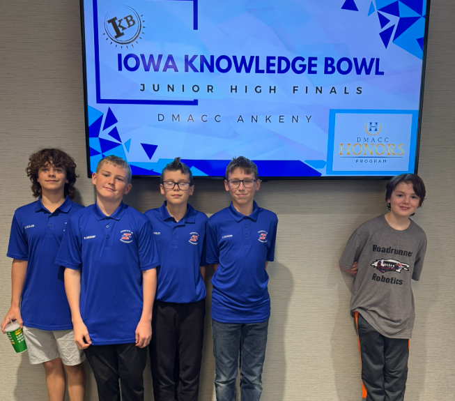 The team of Raph V., Remington H., Lucas F., Blake E., and Ajax B. stand in front of a digital sign reading Iowa Knowledge Bowl Junior High Finals DMACC Ankeny.
