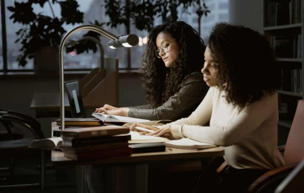 Two young women study by a lamp in a library