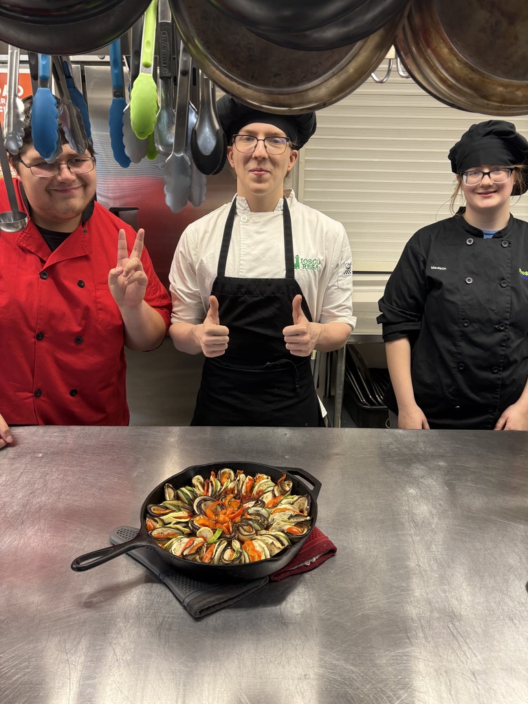 Three students standing behind the counter showing off a skillet of food they made.