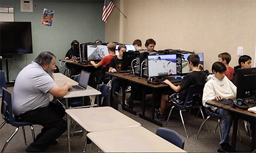 A teacher works on a computer program while students participate in an Esports class