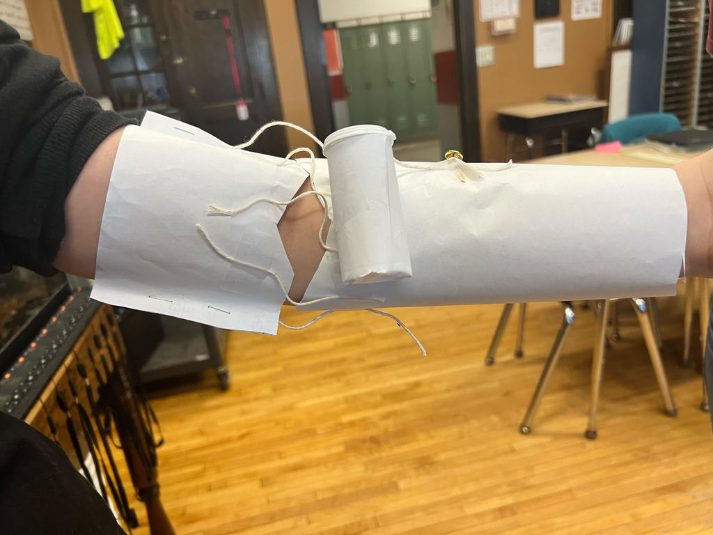 A student's arm is covered with paper. A small cylinder is inside the elbow.