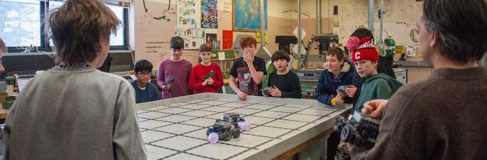A group of students watch a robot race on a table in a classroom setting.