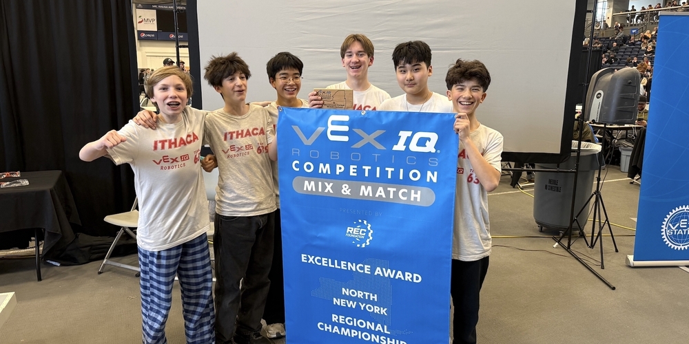 Six middle school students wearing “Ithaca VEX IQ Robotics” team T-shirts stand together at a robotics competition, holding a large blue banner that reads “VEX IQ Robotics Competition – Mix & Match – Excellence Award – North New York Regional Championship 2026.” One student holds a small wooden trophy while the group poses in front of a large projection screen inside a gymnasium filled with spectators and event equipment.