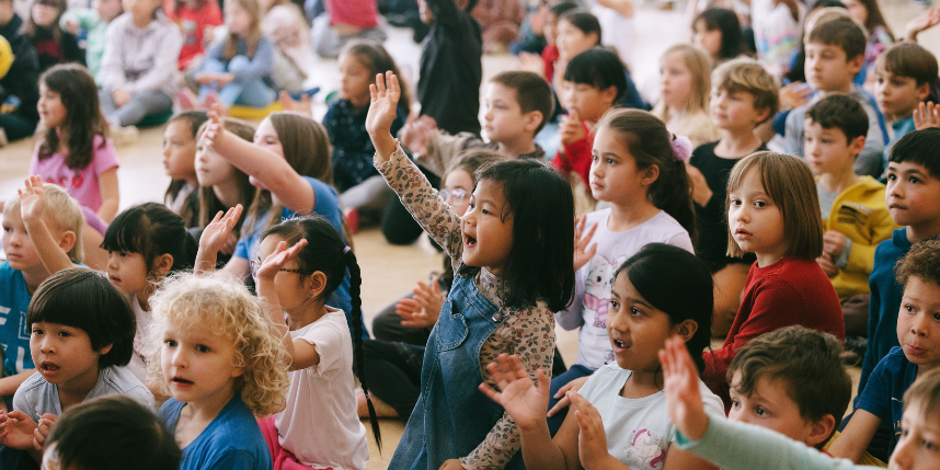 A large group of children sit on a floor and raise their hands; some are clapping.