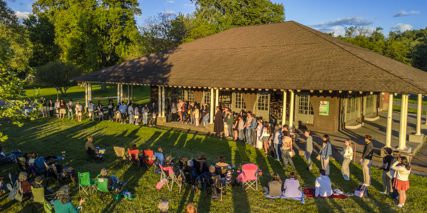 A large group of students stands in a line outside a pavilion at Stewart Park during an evening event. Audience members sit on lawn chairs and blankets on the grass, watching the performance as long shadows stretch across the lawn in the golden sunset light. Trees and blue sky frame the scene in the background.