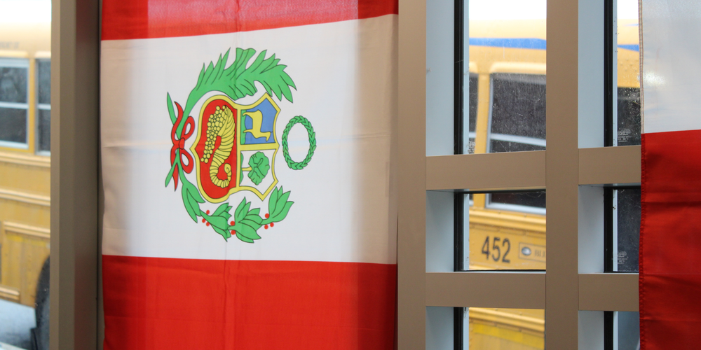 A red and white Peruvian flag with a central coat of arms hangs indoors near a window. Through the window, yellow school buses are visible outside.