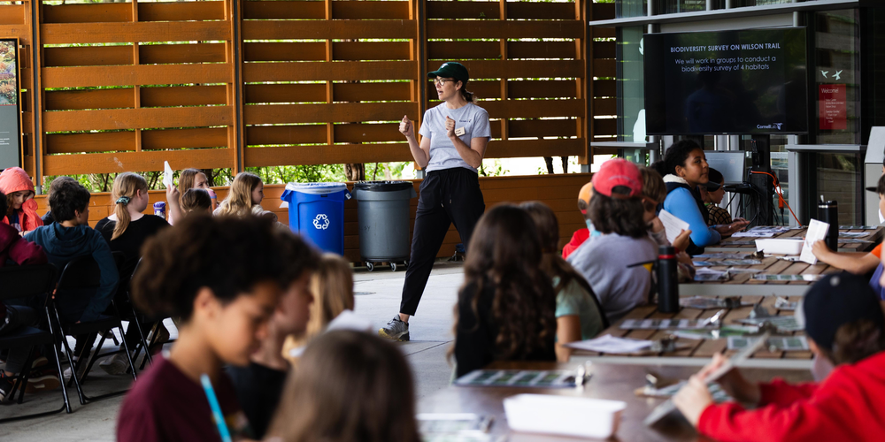 Instructor stands at the front of an outdoor covered classroom, speaking and gesturing to a group of elementary-age students seated at picnic tables. Students are working with papers and clipboards, while a screen behind the instructor displays a lesson on conducting a biodiversity survey.