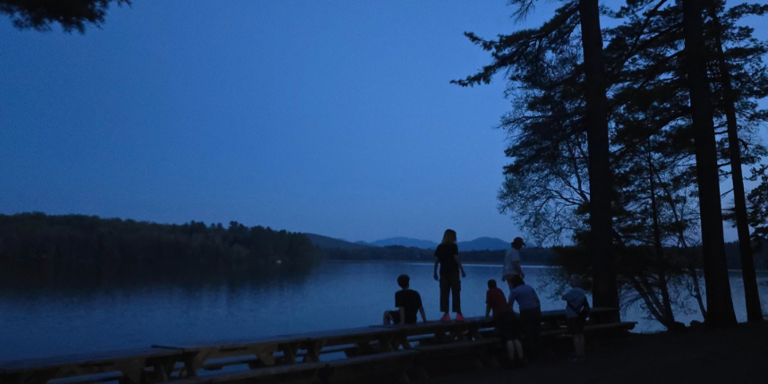 People stand by a lake at dusk, surrounded by trees and mountains in the background.