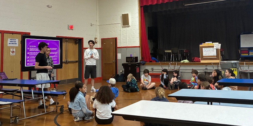 Three IHS students stand at the front of the cafeteria  leading a presentation to a group of south hill students who are seated on the floor in a circle.