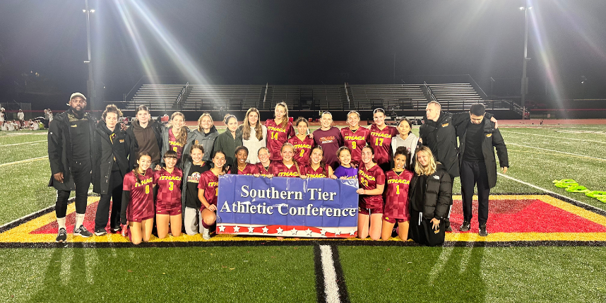 The Ithaca High School girls varsity soccer team poses together on the field at night, smiling and holding a “Southern Tier Athletic Conference” championship banner. The players wear maroon and gold uniforms, and coaches stand alongside them under bright stadium lights.