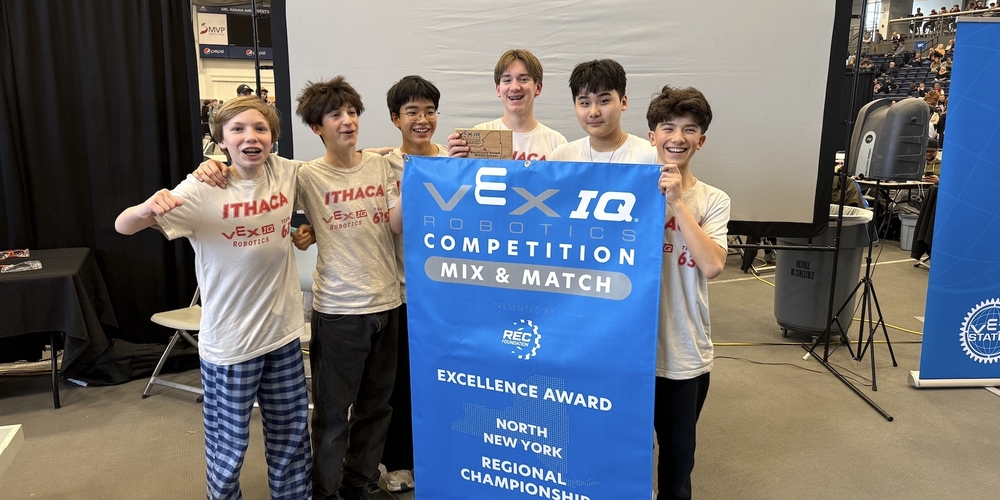 Six middle school students wearing “Ithaca VEX IQ Robotics” team T-shirts stand together at a robotics competition, holding a large blue banner that reads “VEX IQ Robotics Competition – Mix & Match – Excellence Award – North New York Regional Championship 2026.” One student holds a small wooden trophy while the group poses in front of a large projection screen inside a gymnasium filled with spectators and event equipment.