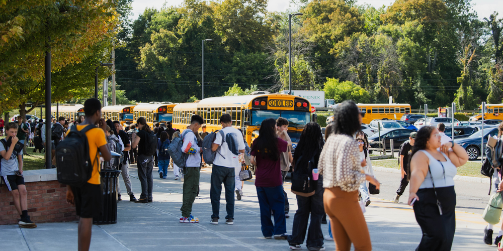 Students walking along a sidewalk at school dismissal with multiple yellow school buses lined up for pickup on a sunny afternoon.