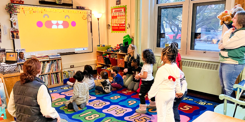 A group of young children sit and stand on a colorful classroom rug watching a bright, smiling cartoon face projected on a large screen. Two adults sit with the children, while another adult stands nearby. The classroom is decorated with books, toys, and learning materials, and sunlight streams in through large windows.