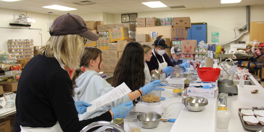 A woman and three girls wearing blue gloves, aprons, and hats work at a long table.