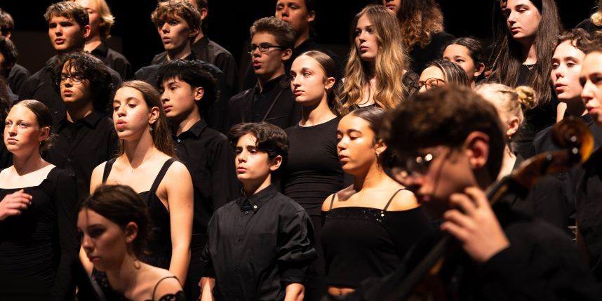 Large group of students on stage dressed in black; one student plays a cello; others stand still.