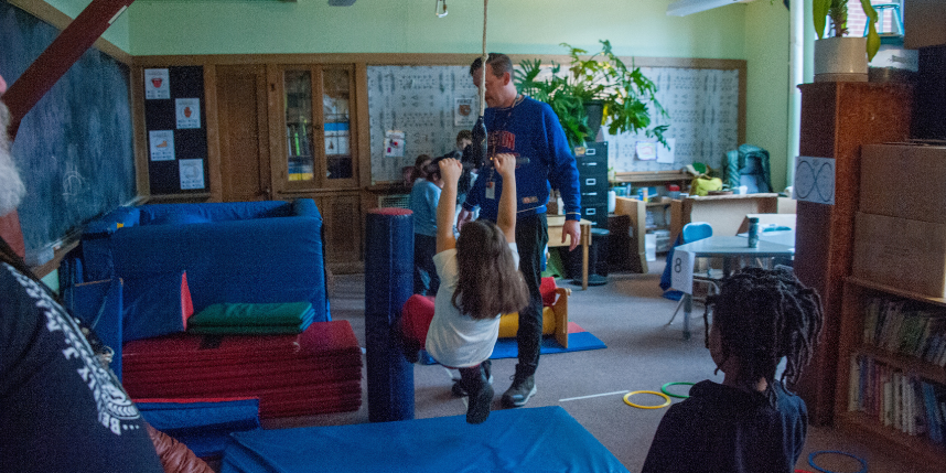 An adult supervises as a child swings from hanging handles over padded mats in a classroom play and movement area, while other children watch nearby.