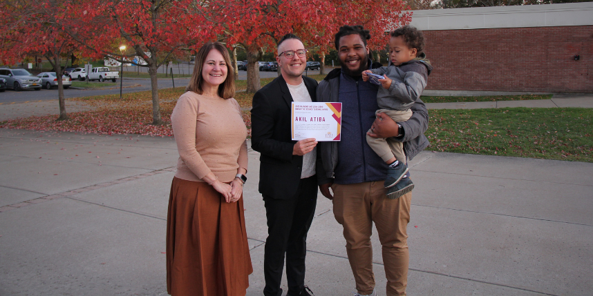 Three adults stand outside on a sidewalk lined with trees with red autumn leaves. The person in the center holds a certificate that reads “Akil Atiba.” The person on the right is holding a small child. All are smiling.