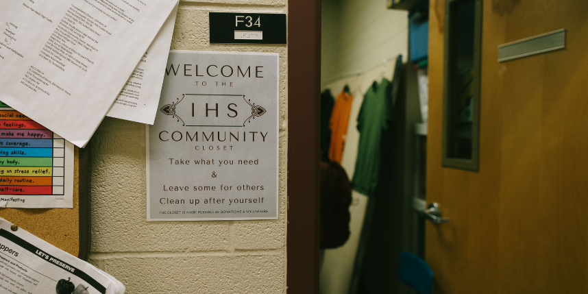A sign on a beige cinderblock wall reads “Welcome to the IHS Community Closet. Take what you need & leave some for others. Clean up after yourself.” The sign is posted outside a room labeled F34. Through the open doorway, clothing items hang on a rack inside the small, softly lit room. Various papers and notices are pinned to a bulletin board on the left side of the image.
