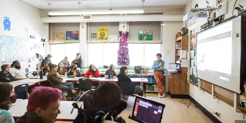 A classroom full of middle- or high-school students sits in groups at tables, facing a large screen where a student at the front of the room is presenting. The presenter stands near a projector and computer setup, holding a microphone. Students and a few adults watch attentively. The room is decorated with posters, artwork, and a purple dress hanging on the wall. Light from large windows brightens the space.